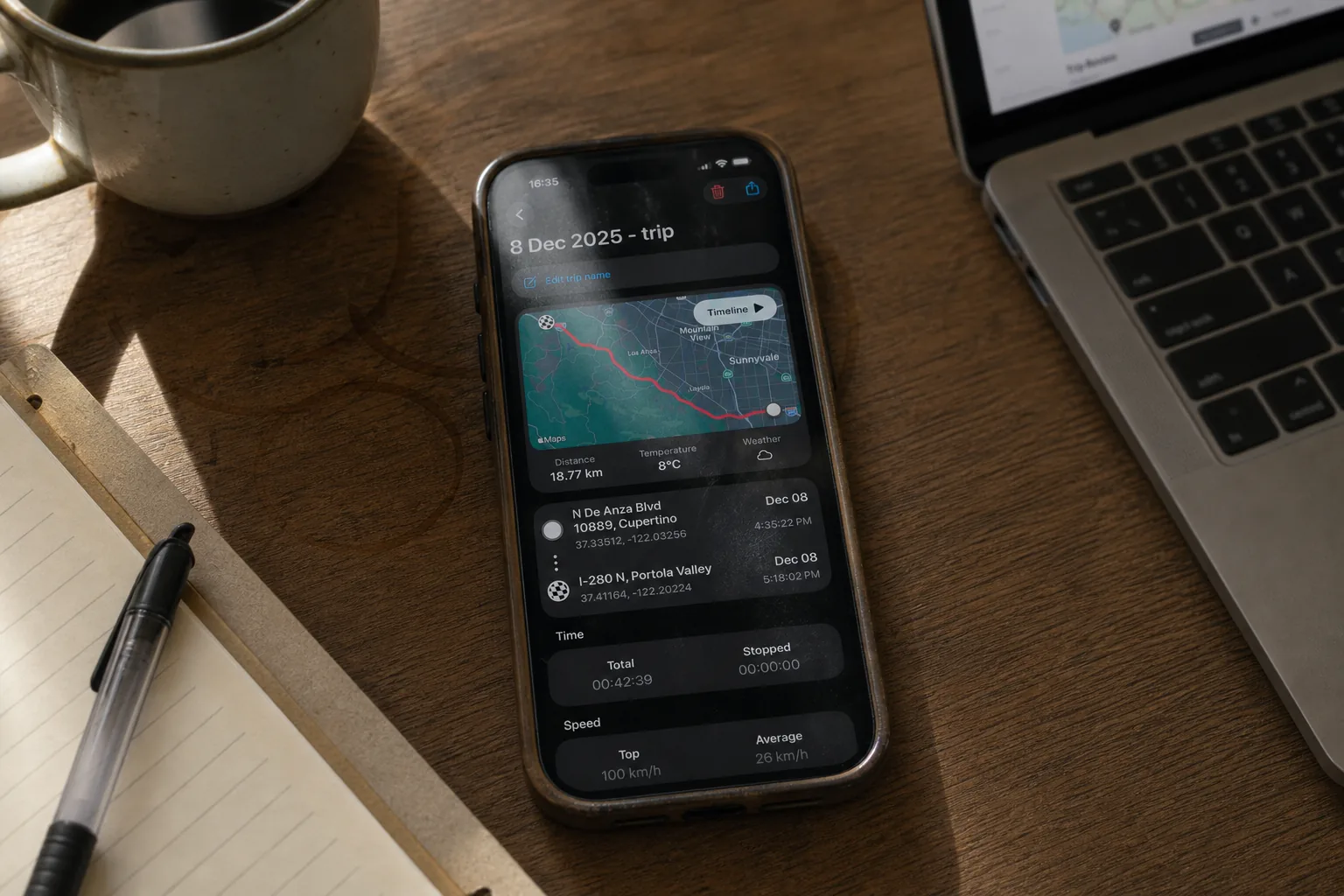 Ein iPhone liegt auf einem Holzschreibtisch neben einer Kaffeetasse, MacBook und Notizbuch — es zeigt die Fahrt-Detailansicht von GPS Speedometer mit Routenkarte und Statistiken