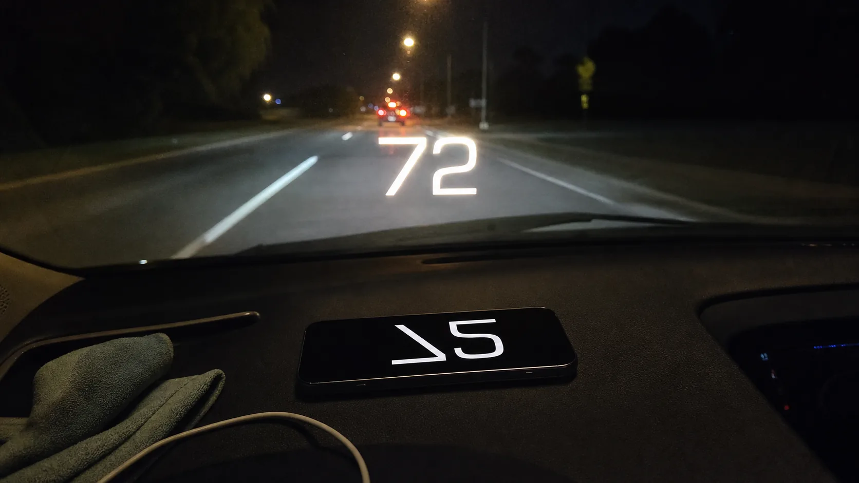 GPS Speedometer's HUD mode in real use — driver's POV at night, iPhone face-up on the dashboard with the speed numeral reflected on the windshield