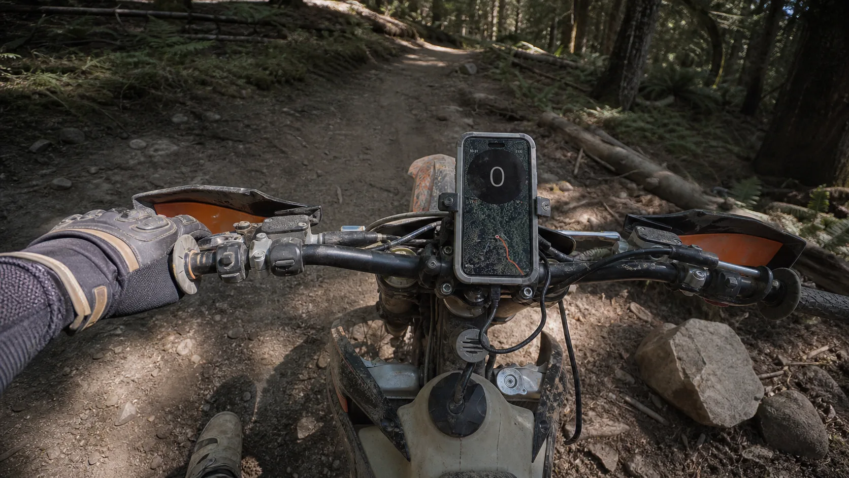 GPS Speedometer running on an iPhone in a heavy-duty rugged case on a dirt-bike handlebar mount, packed-dirt trail extending through forest