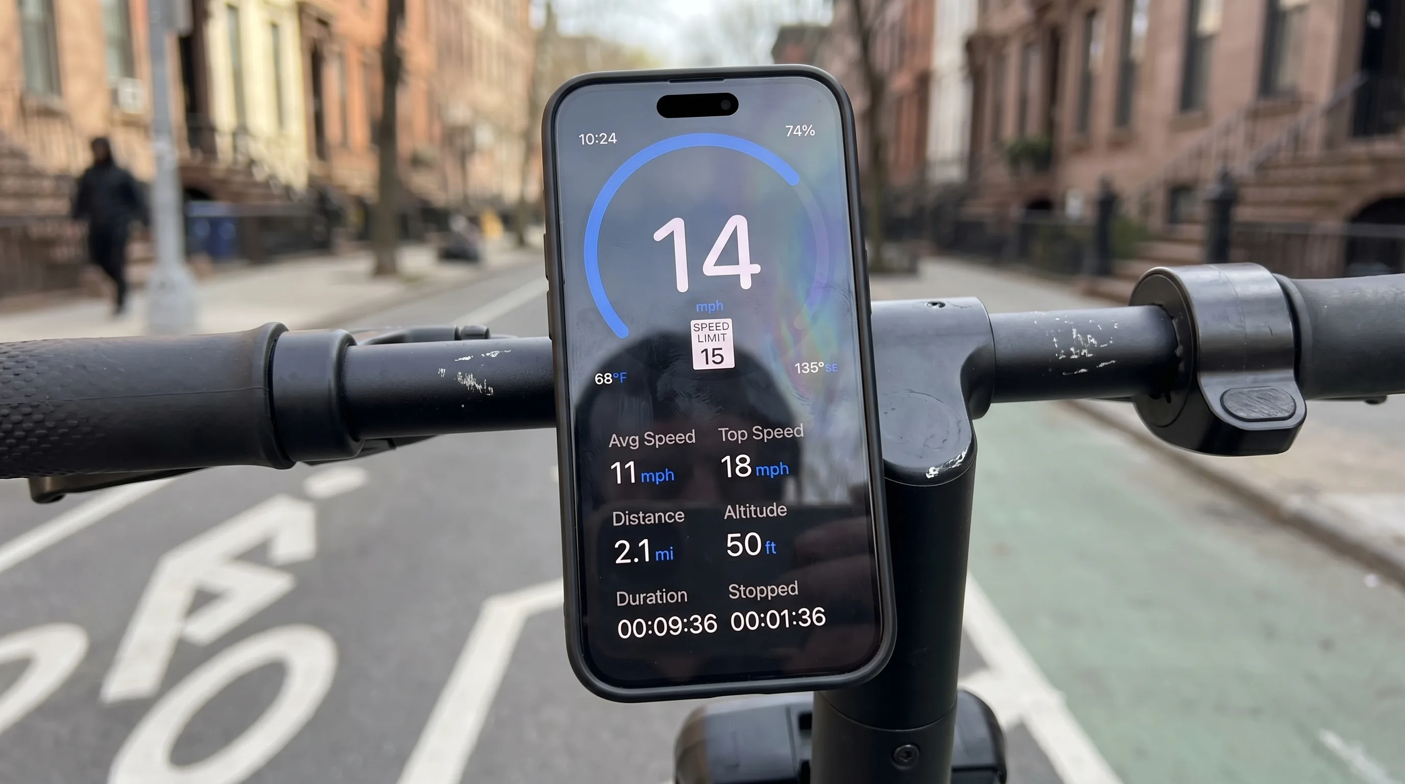 Enge Cockpit-Nahaufnahme eines Steh-E-Rollers auf einem Brooklyner Radweg, iPhone in der Lenkerstangen-Halterung zeigt 14 mph neben der 15-mph-Tempolimit-Anzeige
