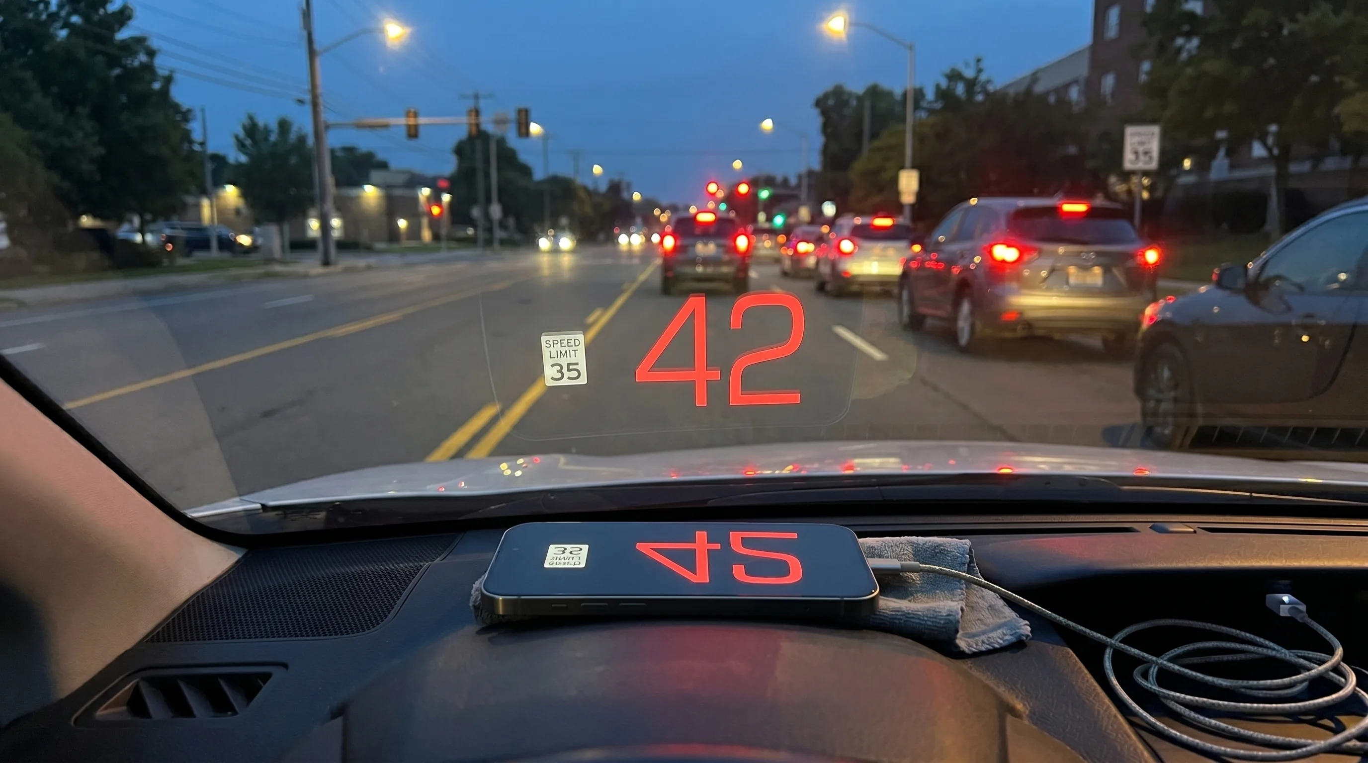 Fahrer-Perspektive in der Dämmerung, mit dem iPhone flach auf dem Armaturenbrett, das die Tempolimit-Warnung auf die Windschutzscheibe spiegelt